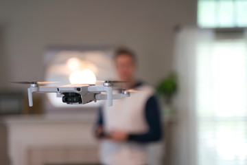 Drone flying indoors with pilot visible in background. Amateur drone flight. User wearing sweatshirt flying drone inside of home on cold day. White drone flying inside.
