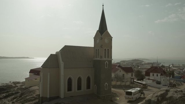 Lockdown shot of church by houses in town against sky, bus on street with buildings near sea on sunny day - Luderitz, Namibia