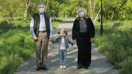 Old grandparents with granddaughter in medical masks walk in park. Coronavirus