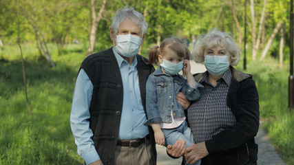 Old grandparents with granddaughter in medical masks walk in park. Coronavirus