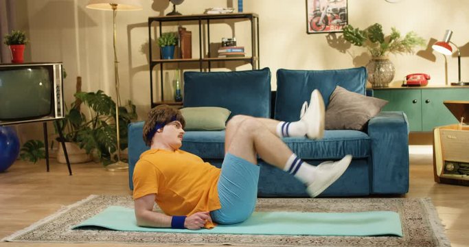 Side view of Caucasian funny retro style athletic man doing laying dawn bicycle on yoga mat indoors. Amusing dark-haired sportsman with mustache working out abs at home near blue sofa. 70s concept