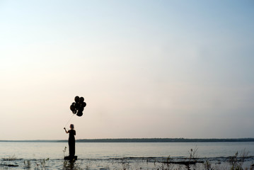 Woman on edge of frame in silhouette stands by the edge of a lake at sunset with black balloons about to let them go letting go of her past