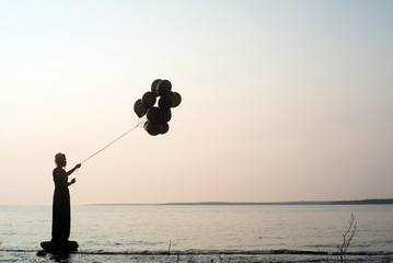 Silhouette of woman holding black balloons at sunset ready to let them go and fly away with lake and water behind her