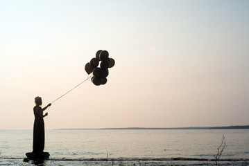 Silhouette of woman holding black balloons at sunset ready to let them go and fly away with lake...