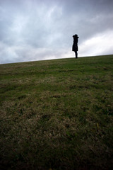 Lone woman wearing black hat and black jacket stands in the distance on top of hill alone and deep in thought against a cloudy sky