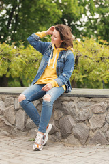 a curly haired young woman is sitting on a stone curb in a city Park in jeans and a bright yellow hoodie smiling with a phone in her hands