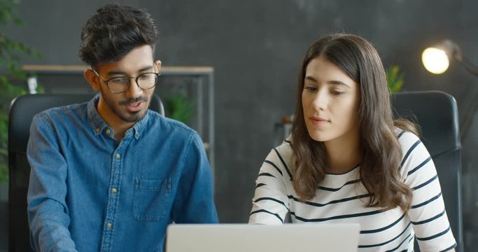 Muliethnic young guy and girl sitting at table together at laptop computer in office and working over new startup project. Mixed-races male and female company workers brainstorming and talking.