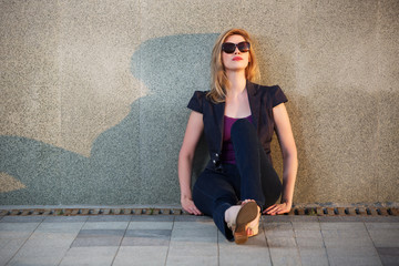 Sad young fashion woman in sunglasses sitting at the wall on sidewalk