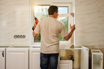 Middle-aged caucasian man stands with his back and holds the kitchen cabinet door and cordless screwdriver, preparing for the process of individual installation in his own house. Quarantine activity