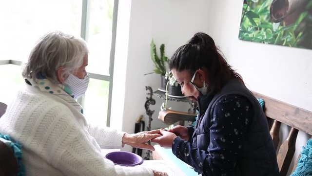 Manicurist making a manicure to a senior client at her home during quarantine both wearing protective face masks