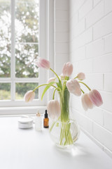Vertical close up of pale pink tulips in glass vase with skincare accessories in background on white table next to window (selective focus)