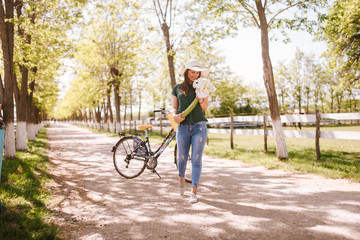 A beautiful young woman with a straw hat, bicycle and flowers is standing in the park. Life in a...