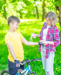 Woman spraying insect repellent or sunscreen lotion on skin her young son, who ride a bike at summer sunny park