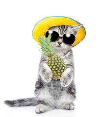 Kitten wearing sunglasses and summer hat holds pineapple and looks at camera. Isolated on white background