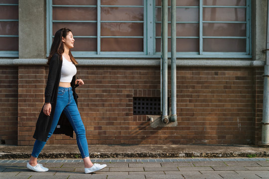 Young trendy asian chinese woman wearing blue skinny jeans and crop top shirts walking on street in summer time. side view full length girl with sunglasses on head walking by red brick wall enjoy sun