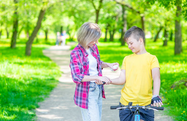 Mom spraying insect repellent or sunscreen lotion on skin her son, who ride a bike at summer park