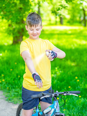 Smiling boy spraying insect repellent or sunscreen lotion on his skin while ride a bike at summer park