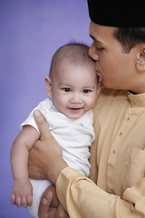 Man in baju melayu and songkok kissing baby on the forehead