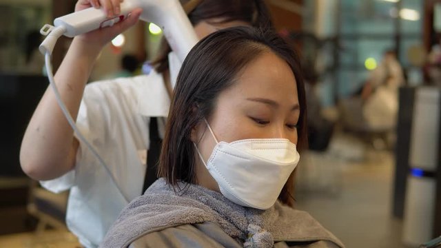 Hairdresser Blow Drying Korean woman's Hair with hairdryer - closeup shot, Korean woman wearing a N95 protective mask, reopen during covid-19