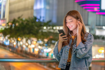 Smiling young beautiful Asian woman listening to broadcast online music from smartphone with earphones in the city at night. Pretty teenage girl enjoying city night light with radio streaming songs.