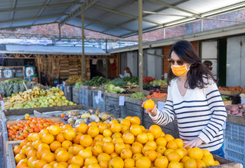 A young white woman with black hair shopping fruits in a chilean street market wearing a protective...
