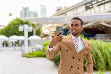 Happy young handsome African businessman taking selfie in the city