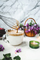 Obraz na płótnie Canvas metal kitchen whisk whips up coffee foam in a large glass Cup with milk on a white marble surface with a burning candle, a wicker basket with a lush bouquet of purple lilacs. dalgona coffee recipe