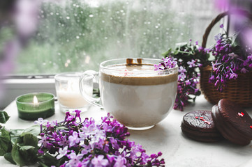 Obraz na płótnie Canvas dalgona coffee with foam, chocolate cookies, burning candles and fresh lush purple-lilac flowers at the wet window with drops and drips from the rain. cozy beautiful Breakfast in bad wet weather