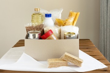 Cardboard box with food on wood table and white napkin. Donation and charity or delivery concept. Oil, canned food, peanut butter, pasta, milk, sugar, rice, buckwheat.