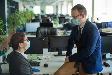 Colleagues in medical masks in an open office space communicate at the desk. A man and a woman in office suits are discussing a working draft. The boss controls the subordinate.