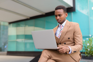 Young handsome African businessman using laptop while sitting outside the building