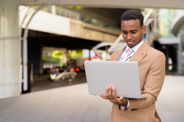 Young handsome African businessman using laptop outside the building