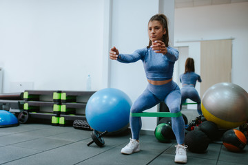 A beautiful young caucasian girl is doing squats with a tape around her knees in the gym