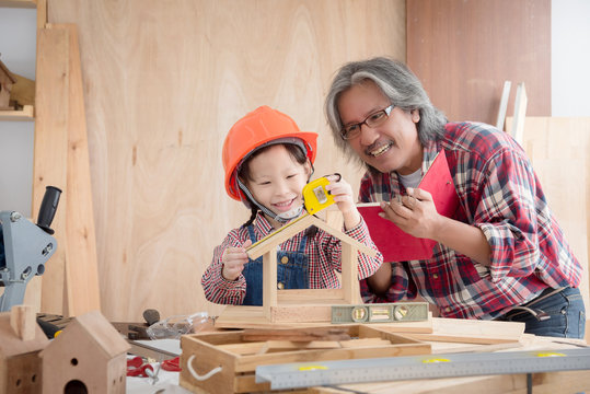 Senior male asian carpenter and grandchild making wooden bird house at home . Little asian girl working at carpentry workshop.