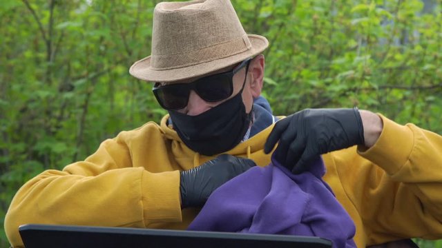 aged man in black mask and sunglasses takes pullovers and compares with laptop screen pictures sitting in garden closeup