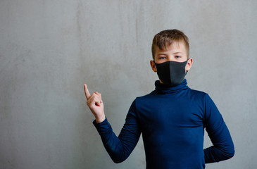 A boy in a black medical mask on his face on a uniform gray background points with his index finger to an empty place next to him and looks at the camera. Place for text