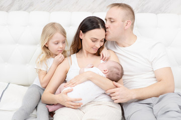 A newborn boy lies in his mother’s arms, a newborn hugs the mother of the newborn and the baby himself, and the older sister sits next to the mother. Happy family concept
