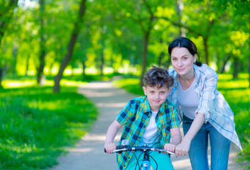 Mom helping her son learn to ride a bike in a summer park. Family weekend breaks. Place for text.