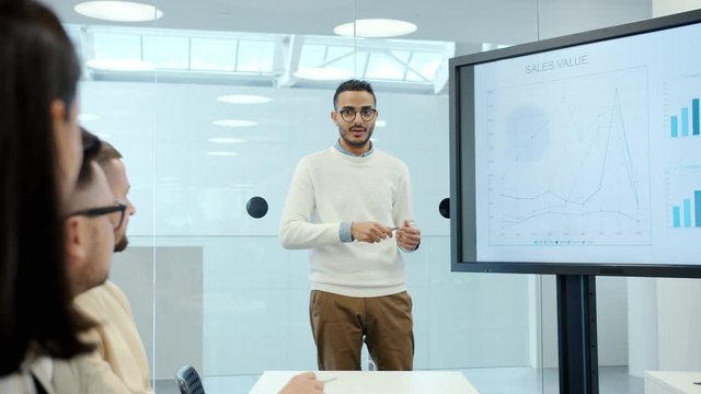 Young Arabian businessman is giving presentation about sales using digital whiteboard in office explaining financial strategies to group of people.