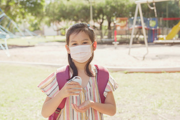 Mixed Asian girl wearing mask and holding hand sanitizer near playground, school reopening, return back to school after covid-19 coronavirus pandemic is over, new normal concept