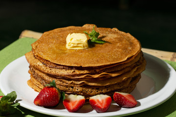 pancakes with strawberries with mint leaves and a slice of butter on a plate, morning homemade breakfast for breakfast, a white dish on a green napkin on a black background