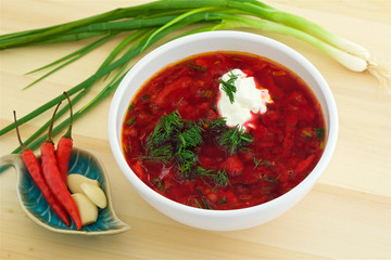 borsch with sour cream and dill in a plate on a wooden table, Russian and Ukrainian traditional soup, green onions in the background, red hot pepper and garlic in a cup
