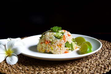 Thai fried rice with dill and lime, a traditional Asian dish, food on a white plate with frangipani flowers on a black background and a straw napkin, rice with vegetables for vegetarians