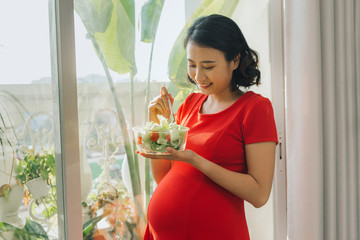 Young pregnant woman eating vegetable salad near window at home