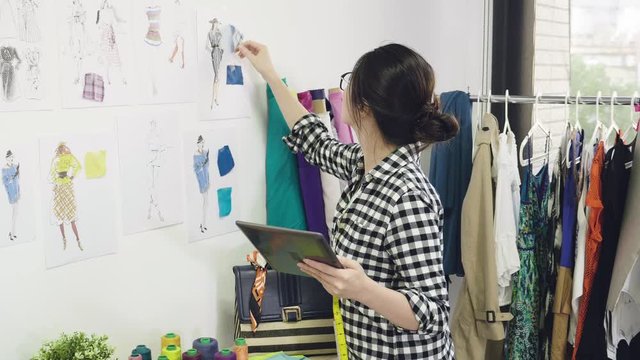 slow motion young female fashion designer standing by wall in office checking fabric on sketches drawings typing on tablet. girl creative dressmaker using digital pad enter information online shop