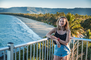 Attractive young girl standing on viewing platform, looking on beach surrounded by mountains and...