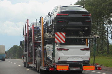 Loaded two-level car carrier truck with two-axle car transporter semi trailer carries new cars on suburban highway road, rear side view close up, automobile delivery