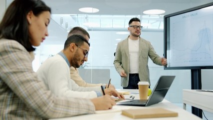 Diverse group of people is listening to business trainer using digital screen during conference, man is speaking about sales and finance in modern office - Powered by Adobe