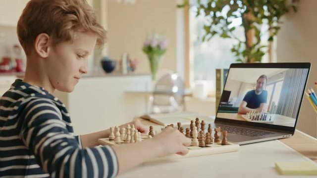 Brilliant Little Boy Playing Chess with His Chess Master, Uses Laptop for Video Call. Remote Online Education, E-Education, Distance Learning, Homeschooling. Over Shoulder