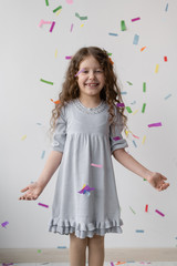 Charming curly brunette in a beautiful dress with her hair is having fun, clapping her hands, playing with multi-colored falling confetti. Happy childhood, a little party.